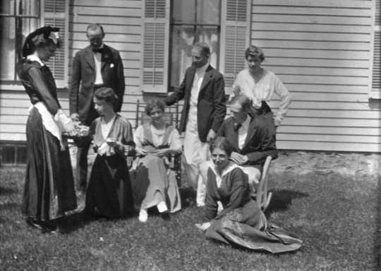 Black and white photograph of Sinclair Lewis and friends, ca. 1916. Lewis sits in the rocking chair at right; his wife, Grace Hegger Lewis, wears a maid’s uniform and stands at left.