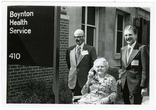 Ruth Boynton with colleagues Don Cowan and Paul Rupprecht at the ceremony where the University Health Service was renamed the Boynton Health Service. She is elderly and sitting in a wheelchair. The two men stand behind her and the sign with the name of the building is in the foreground.